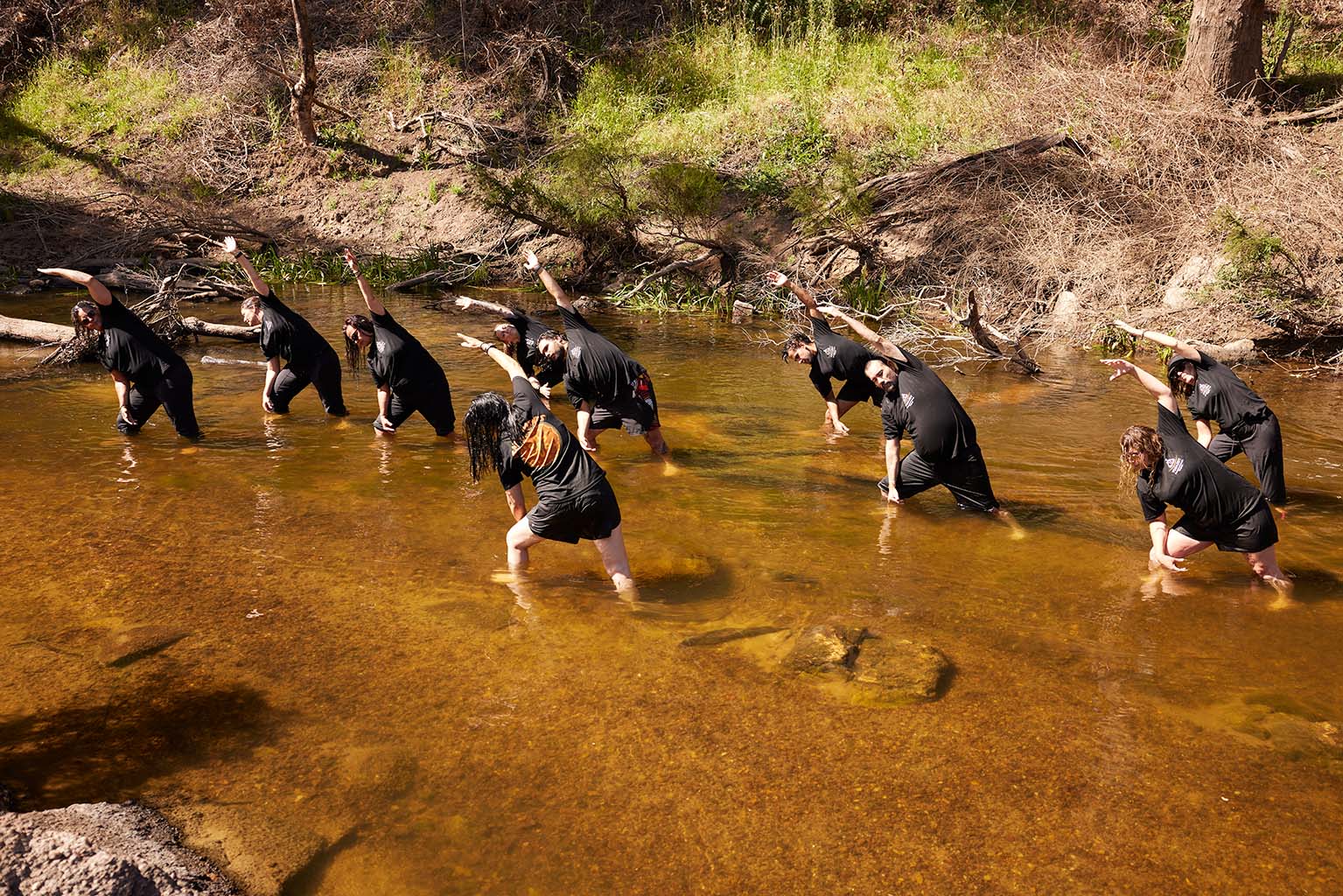 A group shot of all participants facing the facilitator while standing in knee deep water following the facilitators Yoga pose which is bending over to the left with arms outstreched